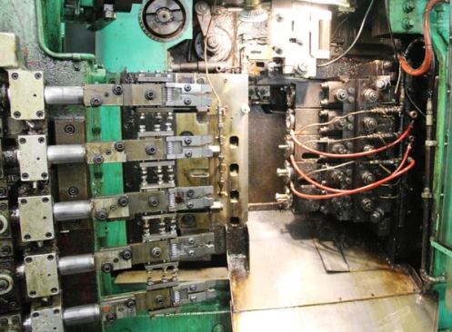 This is the machine from the previous photo with the door open so you can see more of the mechanism. Billets feed in from above and are held in fixtures while dies push in from the right. After each pressing operation the spring-loaded jaws on the left grab the pieces and move them down to the fixture below for the next operation. At the end of this process the roughly formed sockets fall onto a conveyor below.
