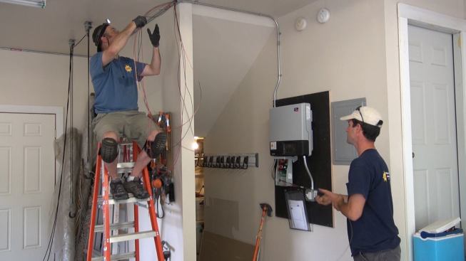 Electrician Ted Houghton and project manager Brandon Bernard pull wire through the final leg of conduit, preparing to complete the circuit from the rooftop panels to the inverter mounted on the wall.