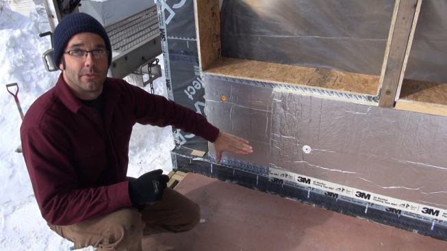 Pollard explains the exterior air barrier and insulation assembly on a freezing cold day in February after a succession of blizzard snowstorms. Visible here are OSB bucks used to extend the window openings; Grace polyethylene and bitumen membrane applied as an air and vapor barrier to the existing board wall sheathing, and integrated into the Grace membrane inserted earlier as a capillary break at the joint between the foundation and the floor frame; a 1.5-inch layer of foil-faced polyiso insulation appied over the air barrier membrane, attached with cap nails (joints sealed with Dow Weathermate tape); and a top layer of 1.5-inch foil-faced polyiso attached with four-inch screws and washers (joints sealed with 3M 8067 flashing tape). Not yet installed here are beveled clapboards in the window sill openings to create sloped drainage, which will be topped with more membrane integrated into the drainage plane formed by the foil-faced foam. “We’re not doing that yet because we don’t want it to get beat up before we put the windows in,” said Pollard.