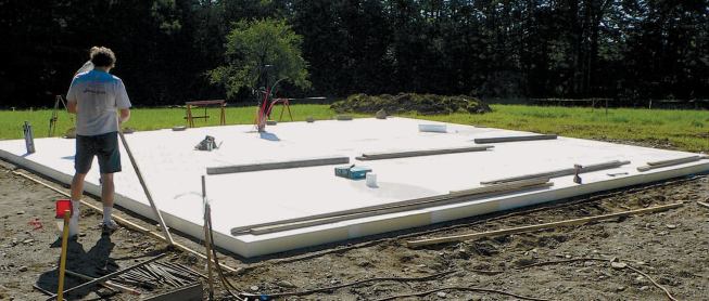 An insulating layer of rigid foam forms a structural base for a monolithic slab foundation. The foam covers the foundation's entire footprint, extending beyond it by about 24 inches on all sides.