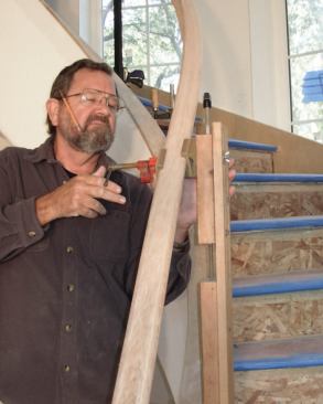 The final piece is a 5-inch-long 1/2-inch pipe clamp attached to the upper 2x4 with a 7-inch-by-3/8-inch carriage bolt, with the nut countersunk into the back of the 2x4. Bending-mold pads protect the rail and allow it to be clamped in place without changing its orientation to the staircase.