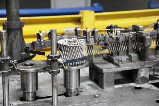 Here’s a closer look at nails being spun into a coil. When the coil is complete the machine lifts it off the spindle and ties it with string prior to boxing. 
Click here for video.