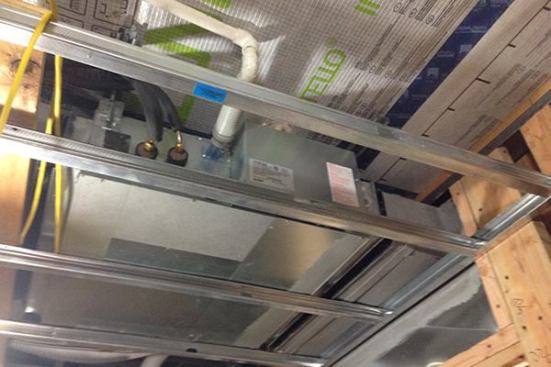 Here’s one of two 9,000-Btu/hr air handlers for the ducted mini-split system, mounted above the ceiling of an upstairs bathroom, directly under the smart vapor barrier that protects insulation in the building’s low-slope roof. Copper refrigerant lines, insulated with foam tubing, sprout from the side of the unit and bend toward the roof (they’ll be attached to the outdoor side of the split system when weather allows that part of the system to be placed). The white piping next to the refrigerant lines is the system’s condensate line (condensate will be pumped under pressure into the home’s plumbing drain system). To the right is a short return plenum that communicates with the home’s hallway.