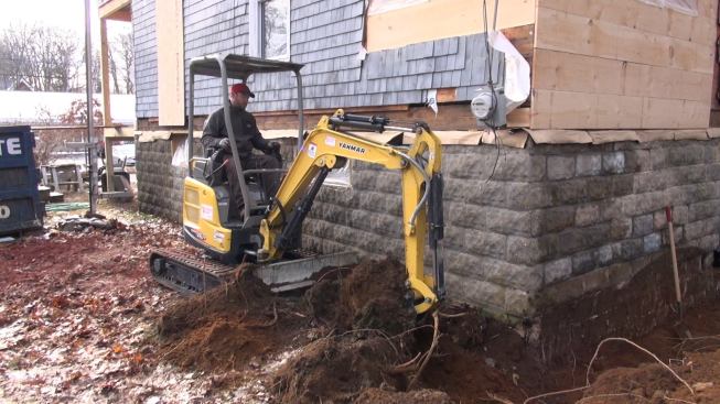 Carpenter Shane Fenton excavates next to the block foundation with a mini-excavator. To place the new foundation under the existing block, the crew will dig down to the level of the lowest course with the excavator, then hand-dig under the wall in four-foot sections to form and place short sections of footing. It will take several days of step-by-step forming and pouring to insert the new continuous footing under the unreinforced block.