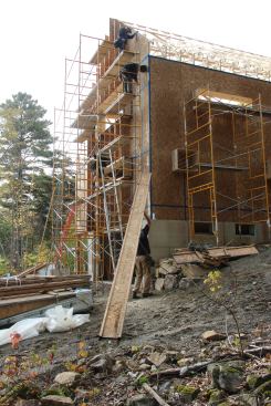 Full-height wood I-joists are applied to the gable end walls before the roof is sheathed with breathable fabric and strapped to create an air space under a plywood nail base for asphalt roofing. (Some houses receive steel roofing instead of plywood and asphalt).