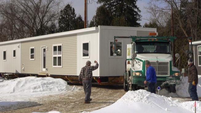 The truck driver and ground guide for Field and Sons (Grafton, New Hampshire) work together to get the rig pulling the heavy modular unit through snowbanks next to the narrow driveway of the mobile home park.