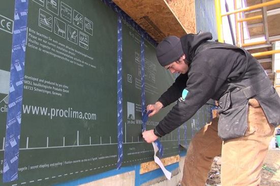 EcoCor carpenter George Reefer applies Pro Clima Tescon tape to the line of staples attaching the DA membrane to the existing sheathing. Staples are located at the wall studs. The tape covers the staples, and will also seal around the fasteners the crew will later use to attach wood I-joists to the walls at the same locations.