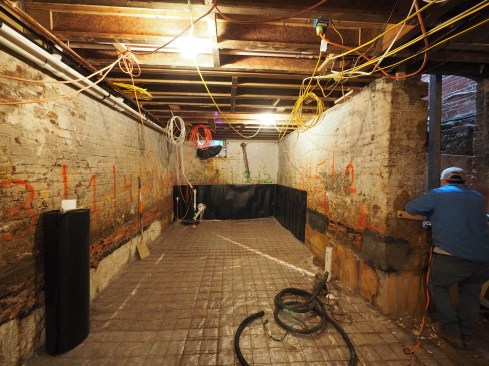 The job has progressed from a state of chaos to order to a semblance of order. Drainage mat is secured to the walls, and the floor is prepped for a slab with a vapor barrier and welded wire.