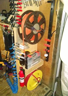Power feeds into the trailer via the 10-gauge cord on the upper reel. The power strip to the right plugs into it. The hose on the yellow reel connects to a “permanently” installed compressor, and provides air for pneumatics used outside the trailer. The box above the hose reel contains collated brads and finish nails.