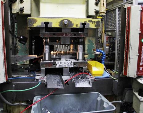 This is looking into the “business end” of one of the smaller Komatsu punch presses. The dies are attached to the flat steel slabs above and below the vertical posts. When the upper die comes down it punches pieces from the strip of steel. The tool part slides down the ramp on the left while the waste comes out the front and lands in the bin in the foreground (
click here for video).