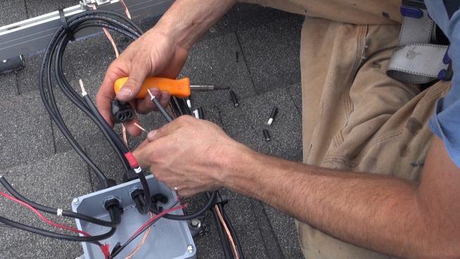 At the junction box on the roof, Bernard connects the home-run cables for the 2 strings of modules to the conductors that feed the inverter on the ground. These wires will be closed into the junction box before the panels are set; current won’t start to flow through this connection until the home-run cables are connected to the panels after all other wiring is complete. (The modules themselves, however, are energized as soon as the sun hits them.)