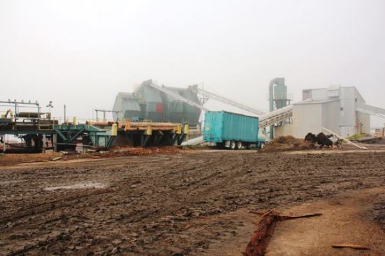 The silver building on the right contains grinding machinery, which grinds all of the bark, sawdust, and offcuts from the mill and makes them into a mulch mix. The mulch is conveyed to overhead bulk bins—the green structure to the left of the truck—and from there dropped into the open top of a semi-trailer.