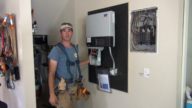 ReVision Energy project manager Brandon Bernard explains the control center of the rooftop solar array his crew is installing. Bernard’s hand is on the direct current disconnect switch, which sits between the high-voltage direct current feed from the roof to the inverter. Above that switch is the inverter itself, which converts the incoming DC power to alternating current and feeds the AC power into the electrical sub-panel at right. In case of a utility power outage, this model of inverter, a transformerless SMA Sunny Boy 4000TL, can also supply the two-plug receptacle mounted below it with as much as 1,500 watts of power (depending on available sunlight). When power from the street fails, however, the inverter will only supply this dedicated outlet—it will not feed any power to rest of the house or back to the grid, for the safety of utility line workers who may be working on downed wires