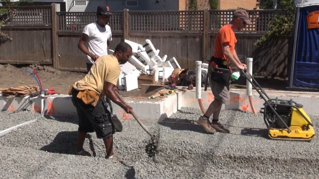 Project manager, Steve Daly, compacts gravel inside the foundation perimeter while coworker Scotland Willis excavates with a shovel for a small section of thickened slab intended as the footing for an interior wall by the stairs of the building.