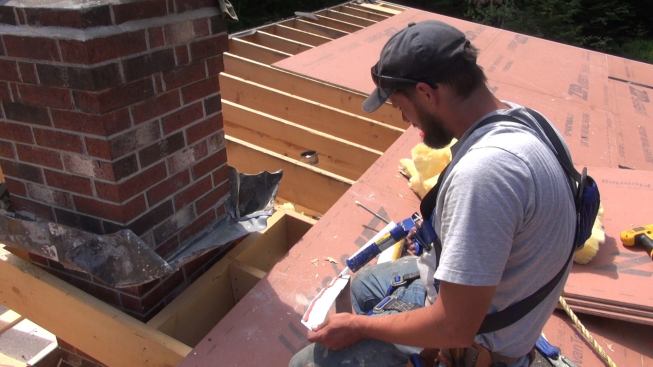 Phillips applies high-temperature sealant caulk to the underside of a piece of flashing before setting the flashing in place. To achieve a good seal, caulking is installed between the metal and the wood framing, as well as at the joint between the metal and the brick chimney.