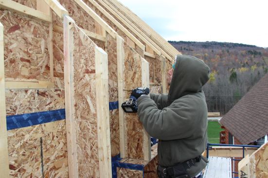 Here, the carpenter screws a short length of I-joist into the wall above the window buck. At the intersection between the walls and the roof, the wall I-joist cavity meets the roof I-joist cavity and allows for continuous insulation over the joint, with only minimal interruption where blocking has been installed.