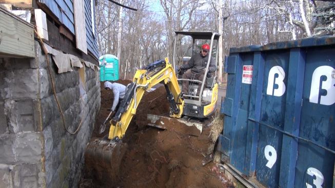 Fenton digs along the base of the wall with the mini-excavator while another crew member hand-digs the last few inches. The ledge on the site is only a foot or two below the lowest course of block in most places, and rises to the base of the wall in several spots.