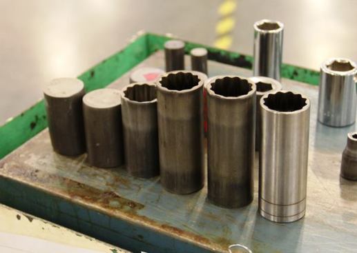 This is what 12 point sockets look like at various stages of forming. It’s important to note that no metal is removed during these particular processes; the change in shape (and length) is due entirely to the displacement of metal. From left to right the pieces are: billet as it enters the machine, after pressing from both ends, after the openings for nuts and bolts have been formed, after the socket is punched deeper, and after the square hole for the ratchet drive has been formed at the opposite end. The shiny piece on the right has been punched and then lightly machined, so it contains slightly less material than the pieces to the left of it.
