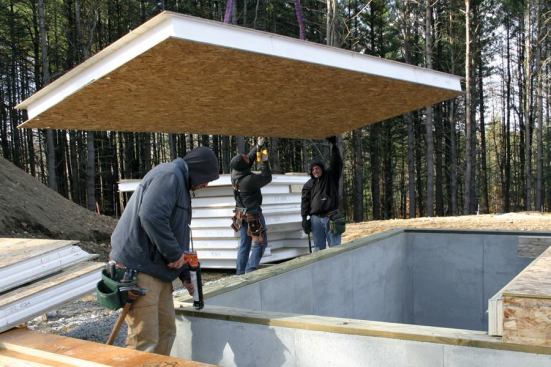 While the floor panel is suspended from the crane, one crew member drills holes at anchor-bolt locations while another applies adhesive sealant to the treated-wood sill.