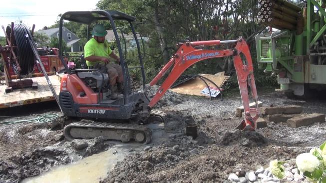 The crew smooths the grade around the well head with the bucket and blade of a skid-steer mini-excavator.