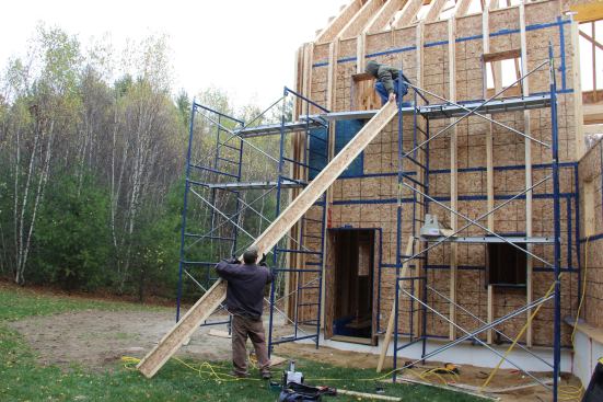 The carpenter on the ground passes a wood I-joist up to the carpenter on the staging.