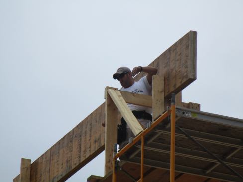 With the LVL carrying ridge beam supported temporarily by scaffolding, a framer gets ready to receive the LVL rafters that will form the site-built supporting truss assembly for the ridge and hips.
