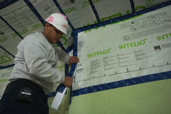 A worker carefully tapes the ProClima Intello membrane to the brick party wall, which has been parged with mortar and coated with Sto Emerald Cote. When the Intello is fully installed and taped to the brick, the dwelling’s occupied space will be fully enclosed within an air control layer consisting of the Intello membrane on the front wall, ceiling, and back wall, and the sealed brick party walls on either side.