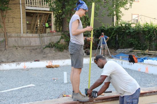 Laura Doyle holds the tape and Scotland Willis screws the angle-iron down to the wood for the screed guide, while Steve Daly sights with the transit to verify that the edge of the angle iron is level. The arrangement is intended to ensure that the slab to be placed will be level to within ? inch.