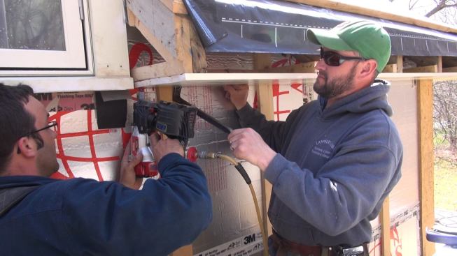 Fenton persuades the tongue-and-groove board for the soffit into position while Pollard pins it in place near the intersection where the eave meets the dormer window.