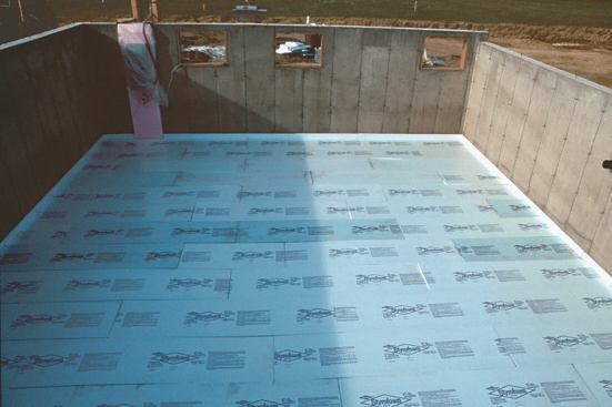 Installing 1 inch of rigid foam insulation under the basement slab keeps the slab warm enough to prevent moisture from condensing on it. The perimeter insulation between the slab and the colder concrete wall provides a thermal break. The cast-in-place pressure-treated window frames will receive low-e argon-filled windows.
