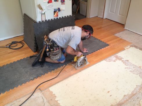 The installers had reversed direction of the hardwood floor in the original kitchen, so the boards needed to be cut back to the blending seam along an adjacent walkway. After removing and saving most of the boards in the original kitchen, Manny rips the board adjacent to the seam so that it can be removed without damaging the walkway boards.