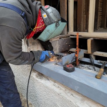 A welder then begins attaching angle-iron cross braces, which will hold the steel legs together and serve as brackets to secure the ends of new floor joists.