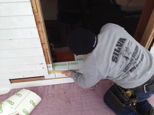 Manny peels the backing off the flashing tape and presses it onto the sloped sill. Using a shim shingle he then gently slips the flap of tape behind the clapboards.