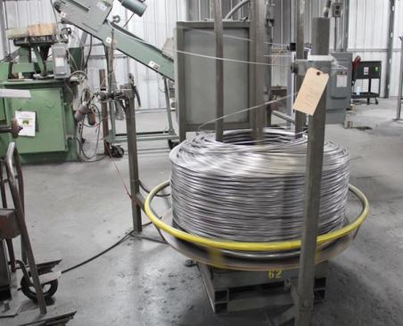 A coil of drawn wire has been placed on a spool at the infeed side of a series of machines that are set up to make coil pallet nails.