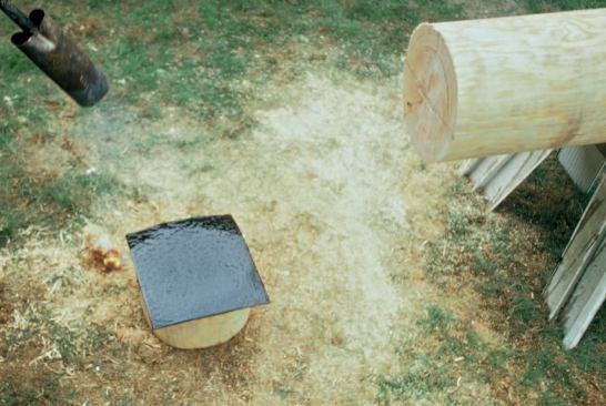 A propane torch heats torch-down roof membrane, used to prevent water from wicking up the log and causing rot. Once heated, the membrane is adhered to the bottom of the log post and trimmed to fit exactly.