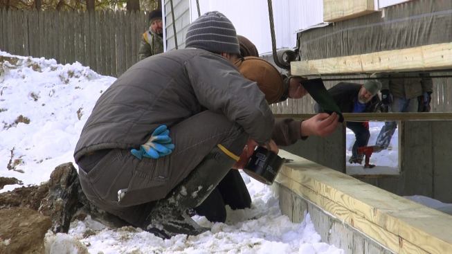 The crew applies bituminous polyethylene vapor barrier tape to the underside of the unit frame before lowering the home onto the treated wood sill.