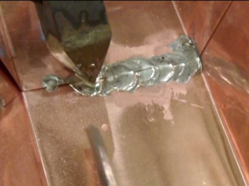 While holding the iron at a shallow angle with the tip against the metal, press the solder stick against one facet of the tip until a drop of melted solder forms, then use the iron to spread the drop across the seam.