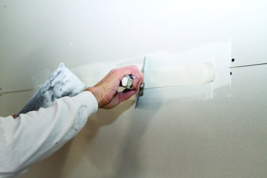 With the tape positioned over the seam, the author draws his taping knife along the length, holding the knife at a steep 60-degree angle while pinning the tape in place with the trowel he uses to carry compound.