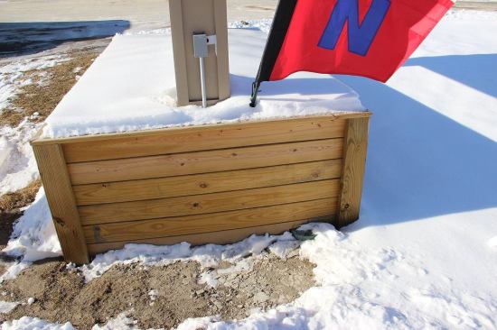 A view of a landscaping detail in Maine: a planting box outside a small office building near a road, filled with soil. This application subjects the wood to conditions that are unusually conducive to decay. But the material, 5/4 treated decking, is not treated to levels appropriate for ground contact, and is not warrantied for this application. Building codes do not regulate planting boxes.
