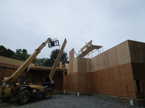 The massive framing members for the hip roof are too heavy to move by hand. Here, an all-terrain forklift raises a wood I-joist rafter up to the roof.