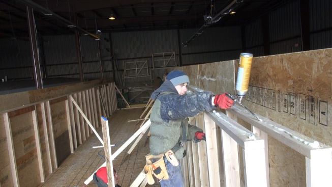An OSB panel serves as the top plate for the bodular unit's double-wall construction, minimizing thermal bridging across this framing juncture. Here, a worker applies a bead of adhesive sealant to the wall plates.