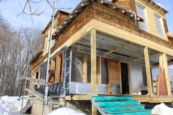 A view of the house during building envelope renovation in mid-winter shows the wall treatment in cutaway view. Existing rough board sheathing and rafter tails are seen at the top of the photo. In the near corner, all the layers of the exterior wall assembly can be seen: first, a layer of Grace Ice and Water Shield, tied into the flap of membrane already inserted between foundation and sill beam; then, successive layers of 1.5-inch foil-faced insulation. Later, the foam will be strapped for a vented rain-screen clapboard siding system.