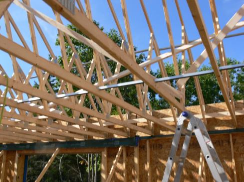 A view of the truss system from below shows how the raised heels of the trusses butt to the OSB skin of the second-floor wall panels. Strips of membrane hanging down from the wall plate will be used to tie the air barrier of the occupied space (created with smart vapor-control fabric), into the wall system air barrier (formed by taped OSB sheathing over the 2x wall frame). In this view you can also see the metal bridging used to space and stiffen the trusses, as well as the diagonal 2x4 truss bracing.