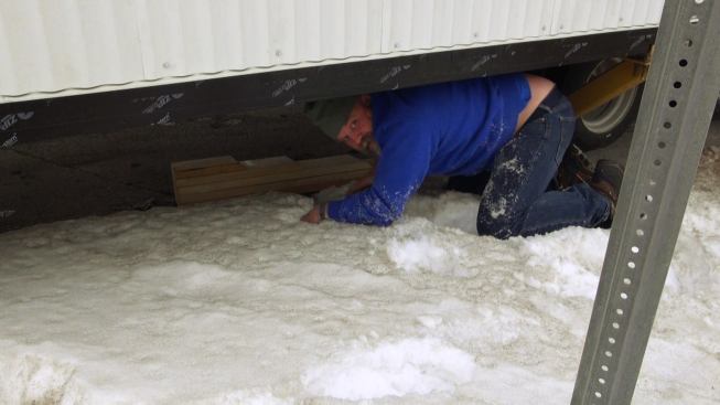 Under time pressure to get his rig through an awkward spot, Steve Davis crawls under the trailer to place a wooden ramp so that the trailer bed can clear an roadside obsttruction.