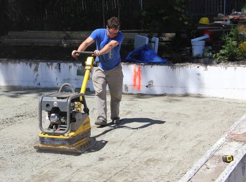 After leveling and compacting the existing soil within the stemwall perimeter, Placetailor’s Evan Smith compacts a layer of engineered fill material containing stone dust along with graded crushed stone, in preparation for adding more gravel to the slab sub-base. Smith explains that without the engineered material, regular gravel would sink into the soft existing soil and be hard to compact, leaving the slab with insufficient support.