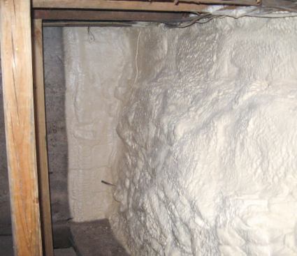 The closed-cell foam on the stone foundation wall at right has been extended a foot or so onto the interior concrete wall at rear to reduce conductive heat loss; the concrete wall was masked before spraying to provide a neat termination. Depending on soils, climate, and other factors, such below-grade applications are sometimes limited to the above-grade portion of the wall — or graduated in thickness, with a thinner layer below grade — to reduce the danger of freezing the footing.