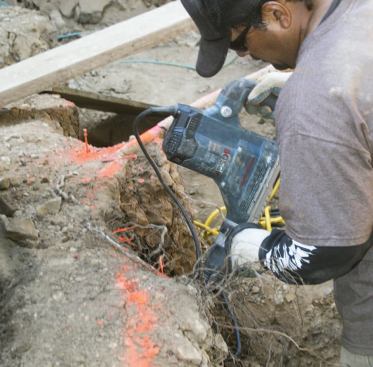 A worker uses a rotary hammer with a spade bit to cut through the hard, gravelly soil.