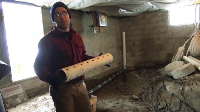 Mark Pollard holds a section of perforated pipe in the basement of the project house, before the slab has been placed. The pipe was used for a radon evacuation system installed in a gravel sub-base beneath the slab. At the rear is the stack for the radon control system, stubbed up. During the blower door test, air flow into the room from the stack stub was readily noticeable until Pollard sealed the pipe with tape.