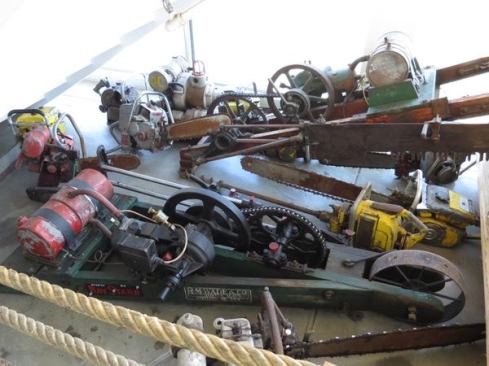 What you want to look at here are the two wooden frames with fuel tanks and motors attached; the one in front is labeled RM Wade & Company. These are drag saws, which came after the hand-operated cross-cut saw but before the chainsaw. They consist of a two-stroke engine attached to a cross-cut saw blade. The one in back has a blade on it; the one in front does not. The blade on a drag saw could be oriented to fell standing trees or cut downed trees into shorter pieces. It’s a clever design, and as hard as it must have been to haul these things into the forest, it had to have been much easier than sawing by hand. To see one in action, search for “drag saw” on YouTube or 
click the link to this video.