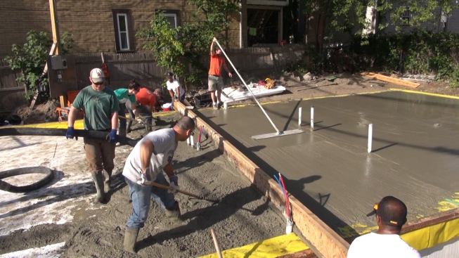 On the morning of the slab pour, three workers shovel and rake concrete while two operate the long screed to strike the concrete level, one handles the hose from the pump truck, and one bull-floats the concrete.